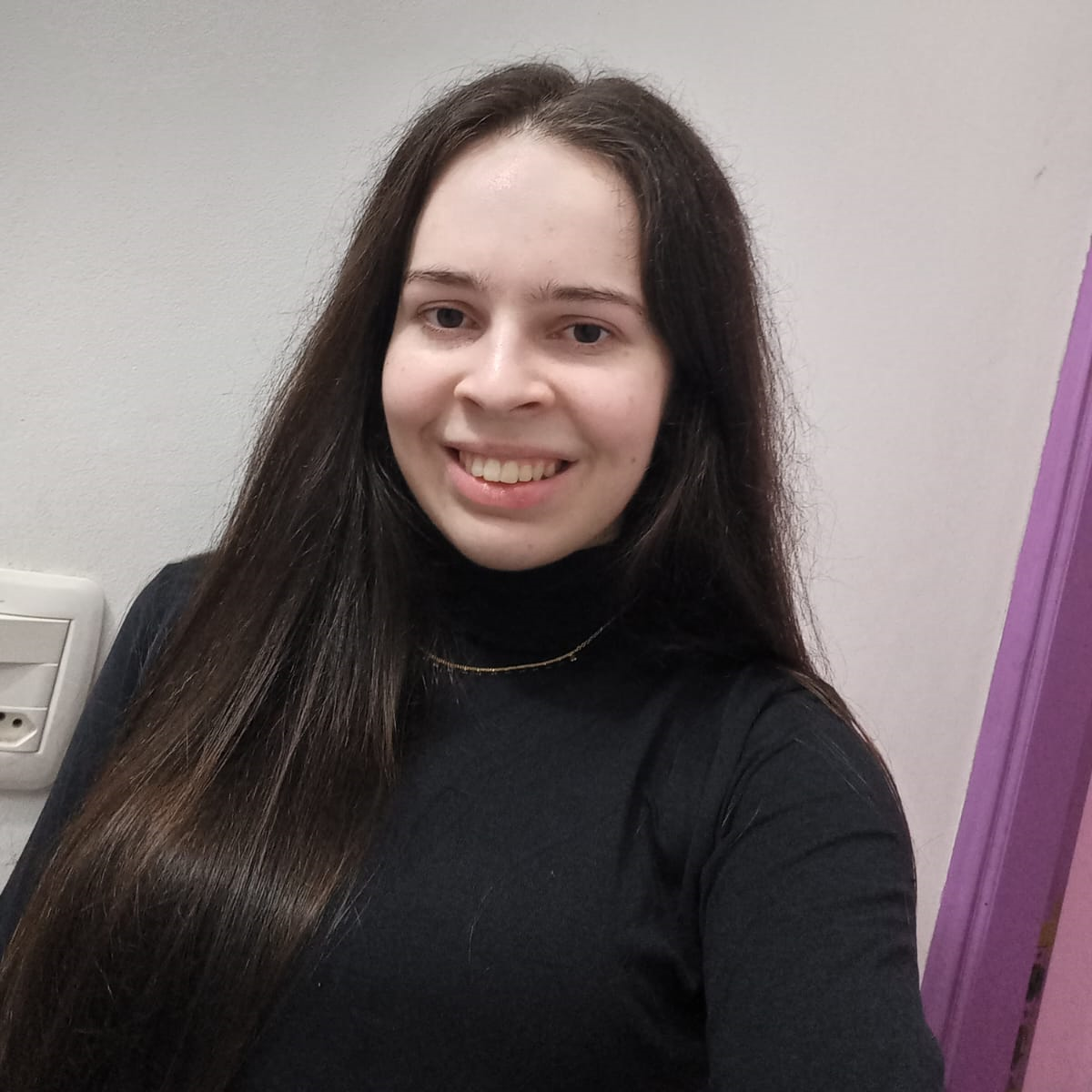 Foto de uma mulher com cabelos longos de cor castanho escuro, pele branca, sorrindo com blusa de manga longa preta e um colar dourado. 